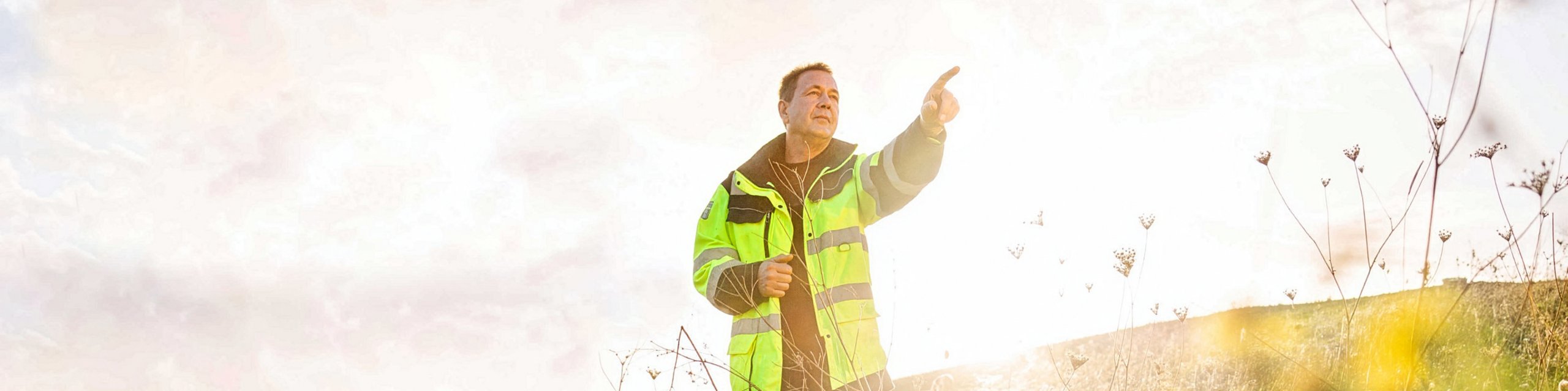 Mann mit Arbeitsjacke steht auf einem Feld mit Sonne im Hintergrund und zeigt in die Ferne